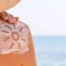 Eine Person mit einem Sonnenhut trägt am Strand Sonnencreme auf die Schulter auf, mit einem Sonnensymbol – zur Vorbeugung gegen Sonnenbrand.